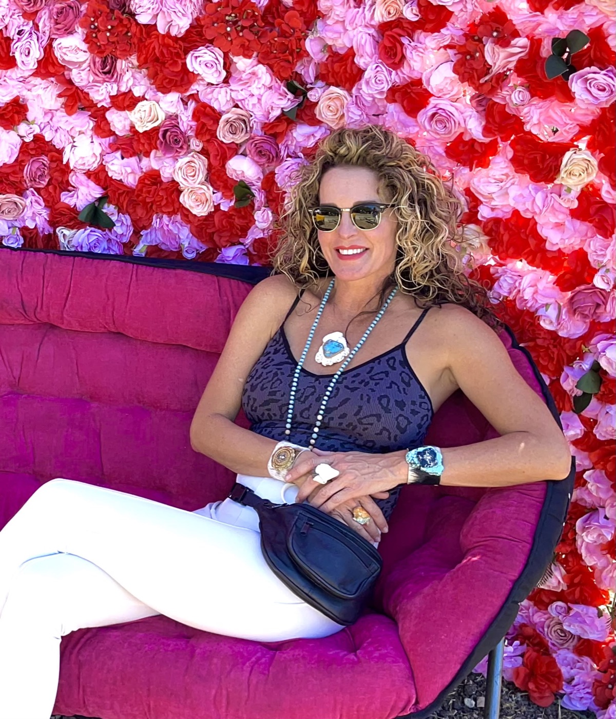 Stephanie Hawley in sunglasses on a magenta sofa against a rose wall, wearing turquoise pendant, cuffs and rings